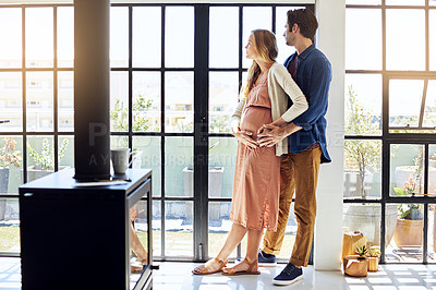 Buy stock photo Shot of a young man embracing his pregnant wife and looking out of a window at home