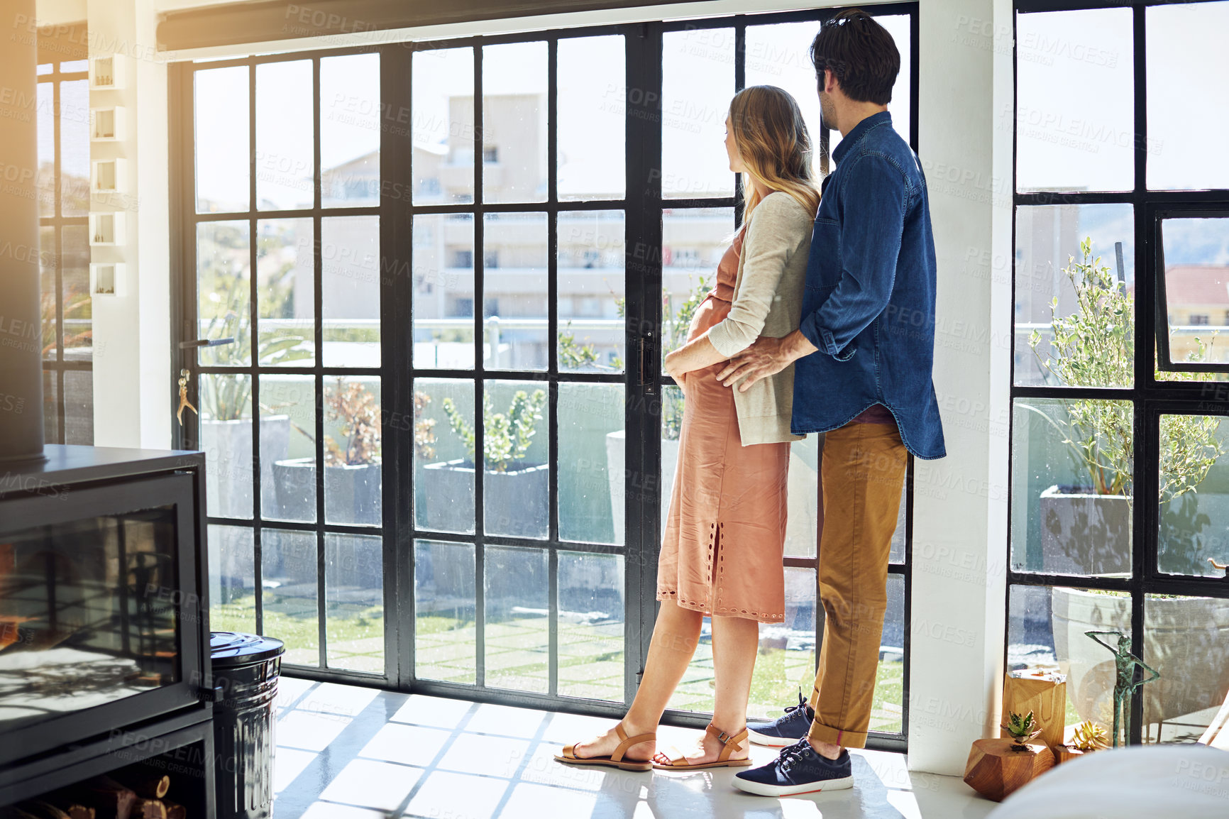 Buy stock photo Shot of a young man embracing his pregnant wife and looking out of a window at home