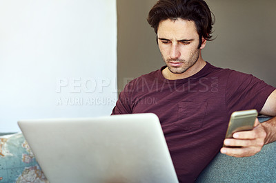 Buy stock photo Shot of a handsome young man using his laptop and mobile phone on the sofa at home