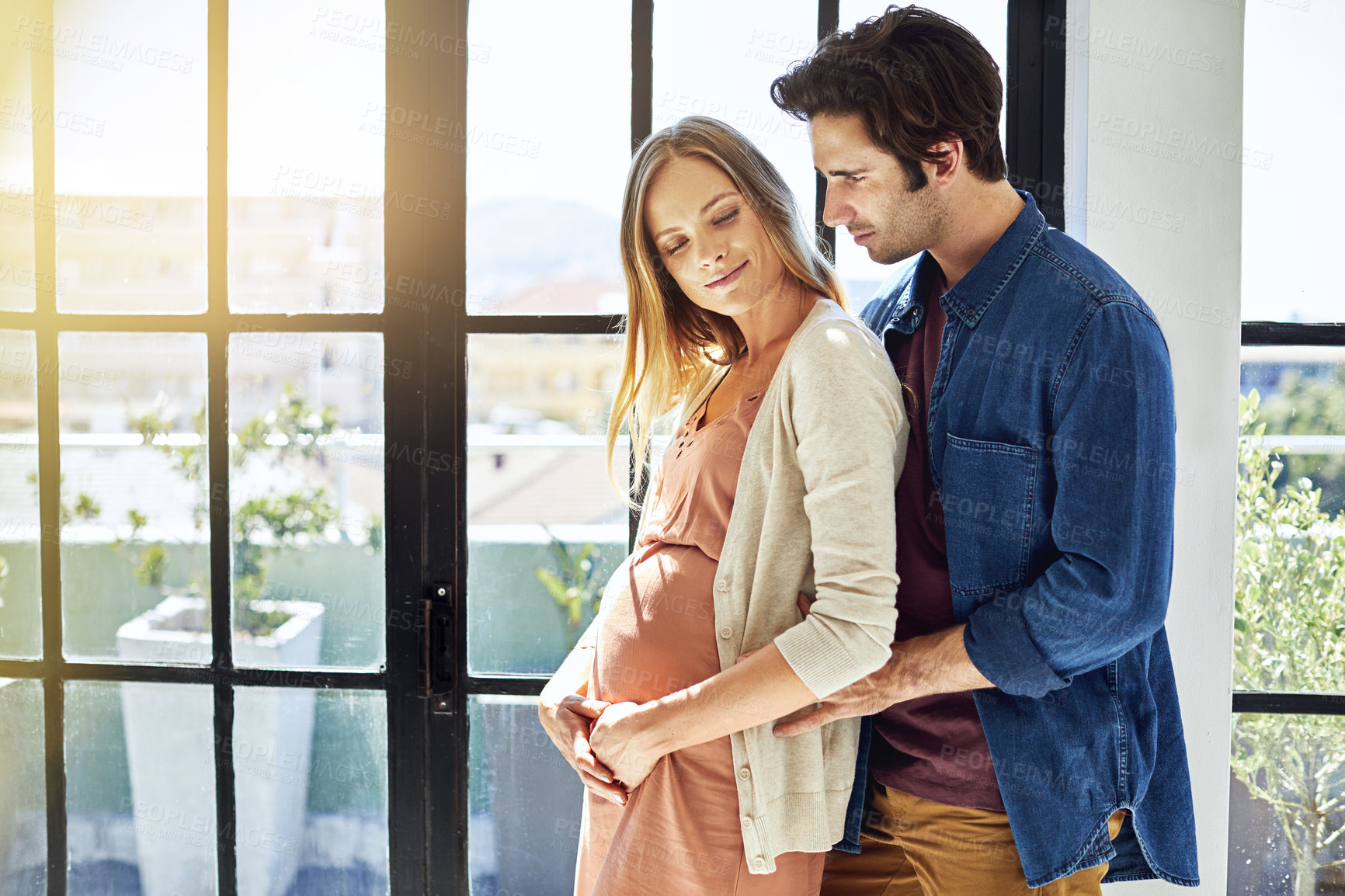 Buy stock photo Pregnancy, happy and couple hug by window for commitment, maternity support and love in home. Marriage, flare and man with pregnant woman for embrace, affection and care for relationship and bonding