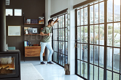 Buy stock photo Shot of a handsome young man having coffee while standing by a glass door at home