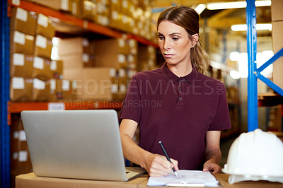 Buy stock photo Shot of a young woman writing notes while using a laptop in a warehouse