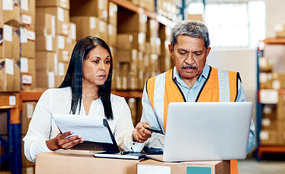 Buy stock photo Shot of a mature man and woman going through paperwork while using a laptop in a warehouse