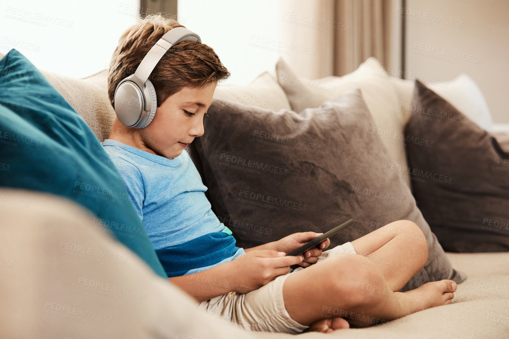 Buy stock photo Shot of a cute little boy using a digital tablet with headphones on the sofa at home