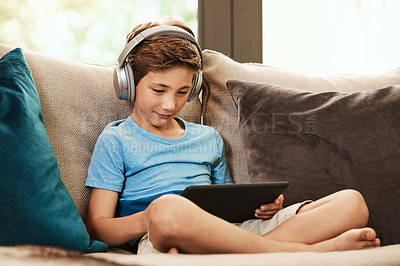 Buy stock photo Shot of a cute little boy using a digital tablet with headphones on the sofa at home