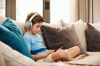 Buy stock photo Shot of a cute little boy using a digital tablet with headphones on the sofa at home