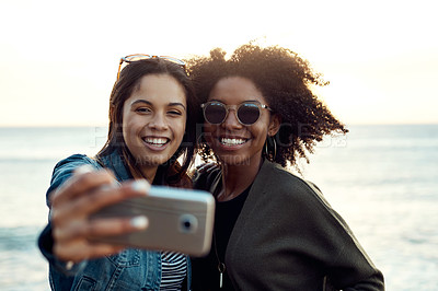 Buy stock photo Shot of two happy young friends taking selfies on a vacation along the coast