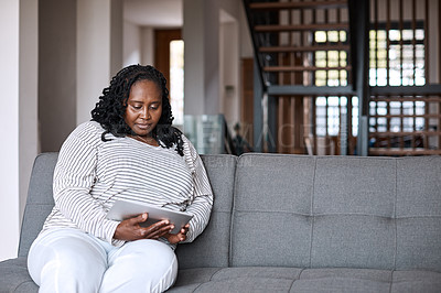 Buy stock photo Shot of an attractive mature woman using a digital tablet while relaxing on a sofa at home