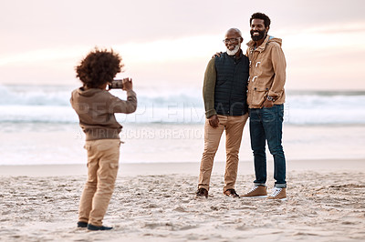 Buy stock photo Shot of an adorable little boy taking a photo of his father and grandfather with a smartphone at the beach