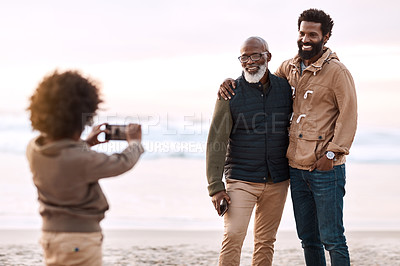 Buy stock photo Shot of an adorable little boy taking a photo of his father and grandfather with a smartphone at the beach