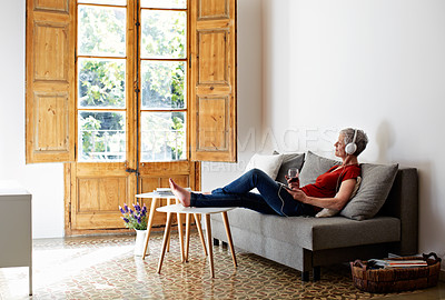Buy stock photo Shot of a mature woman sitting on her sofa listening to music on a digital tablet and drinking wine