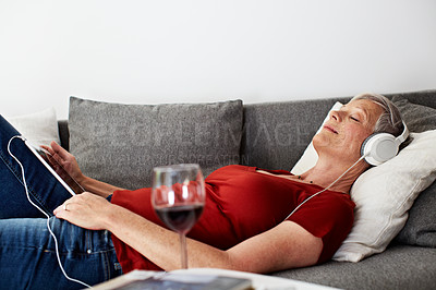 Buy stock photo Shot of a mature woman lying on her sofa listening to music on a digital tablet and drinking wine