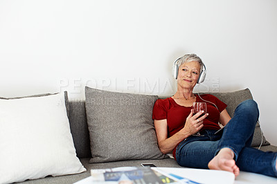 Buy stock photo Shot of a mature woman sitting on her sofa listening to music on a digital tablet and drinking wine
