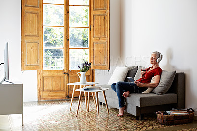 Buy stock photo Shot of a mature woman sitting on her sofa listening to music on a digital tablet and drinking wine