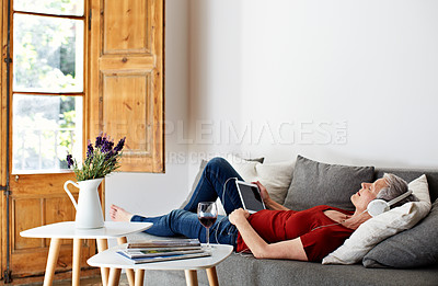 Buy stock photo Shot of a mature woman lying on her sofa listening to music on a digital tablet and drinking wine