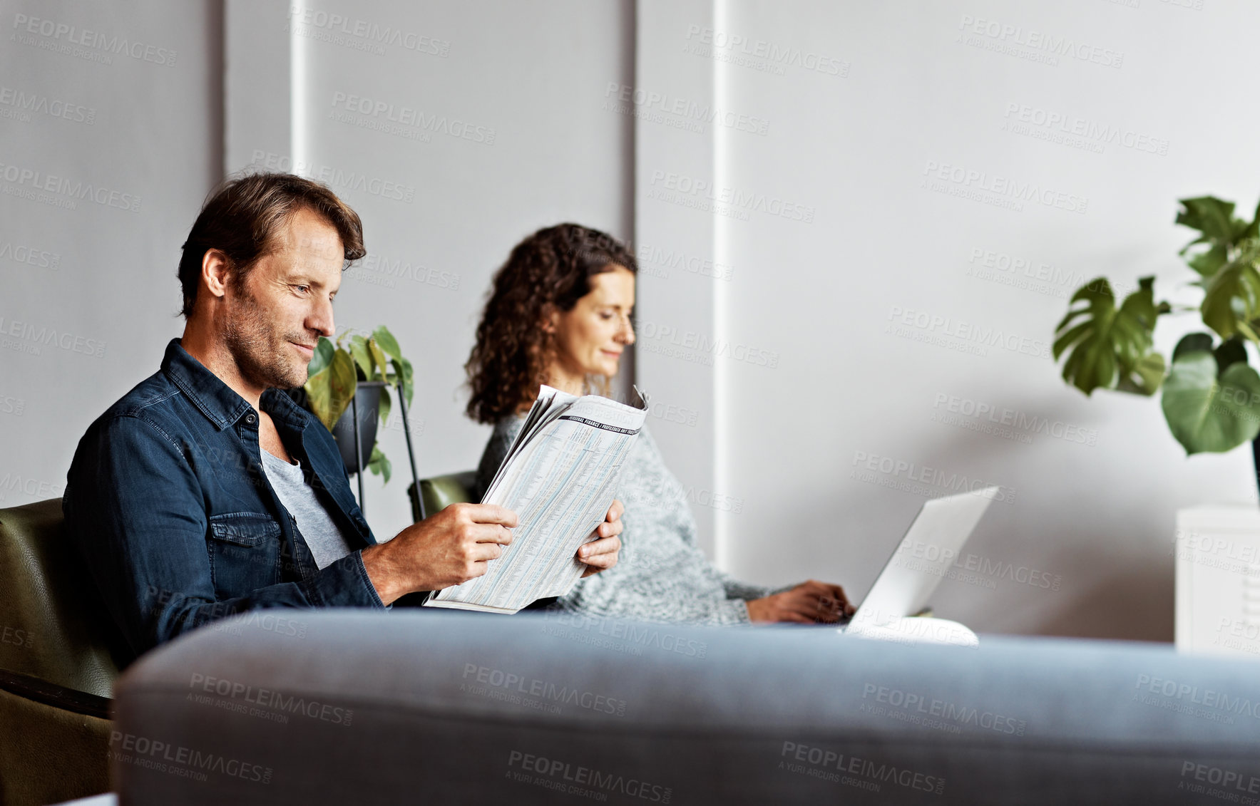 Buy stock photo Shot of a content mature couple sitting in their living room reading the newspaper and using a laptop