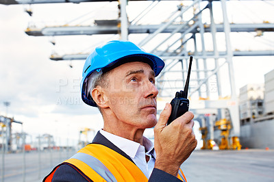 Buy stock photo Shot of a man in workwear standing outside on a large commercial dock talking on a walkie talking