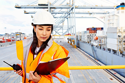 Buy stock photo Shot of a woman in workwear holding a clipboard and walkie talkie while standing on a large commercial dock