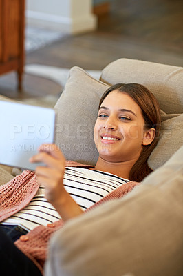 Buy stock photo Shot of a young woman lying on her sofa using a digital tablet