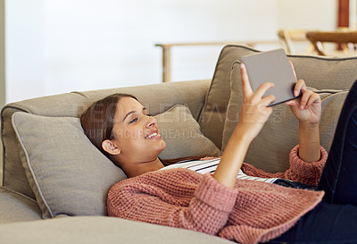 Buy stock photo Shot of a young woman lying on her sofa using a digital tablet