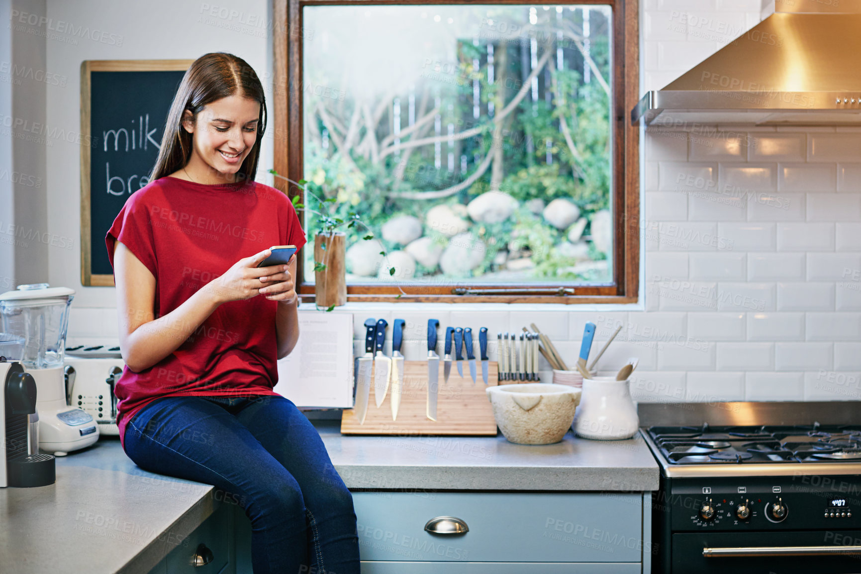 Buy stock photo Shot of a young woman sitting on her kitchen counter using a cellphone