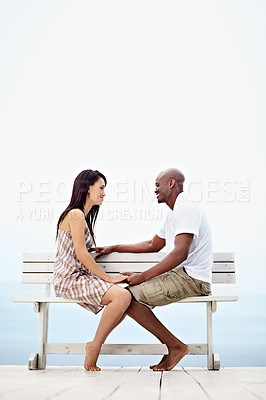 Buy stock photo Shot of a young couple talking together while sitting on a bench by the ocean
