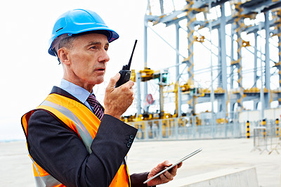 Buy stock photo Shot of a man in workwear standing outside on a large commercial dock talking on a walkie talking