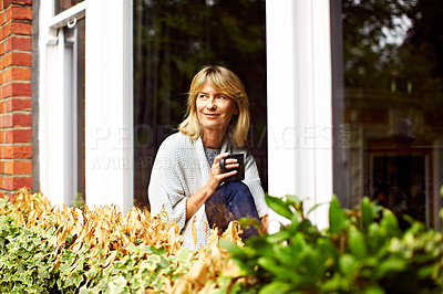 Buy stock photo Shot of a mature woman drinking coffee while looking out of her living room window