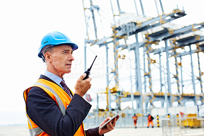 Buy stock photo Shot of a man in workwear standing outside on a large commercial dock talking on a walkie talkie