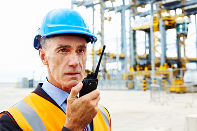 Buy stock photo Shot of a man in workwear standing outside on a large commercial dock talking on a walkie talkie