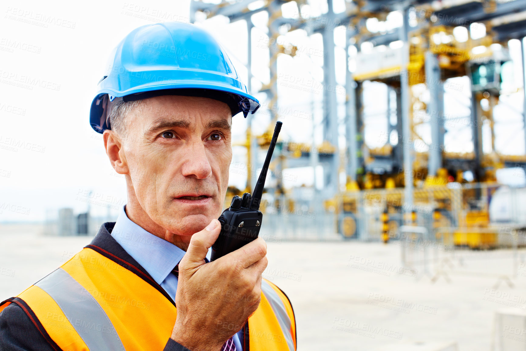 Buy stock photo Shot of a man in workwear standing outside on a large commercial dock talking on a walkie talkie