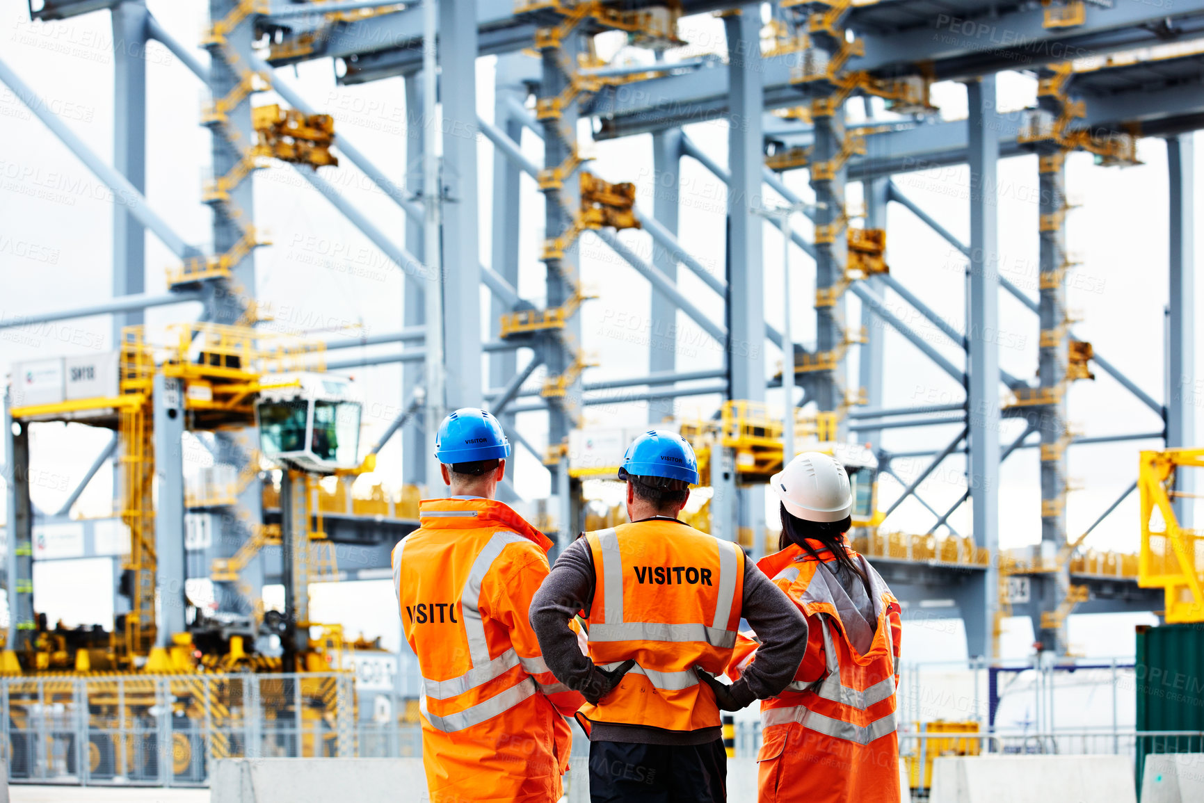 Buy stock photo Rearview shot of of three dock workers looking up at loading cranes