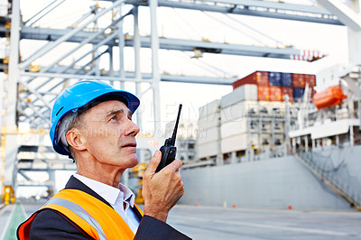 Buy stock photo Shot of a man in workwear standing outside on a large commercial dock talking on a walkie talkie