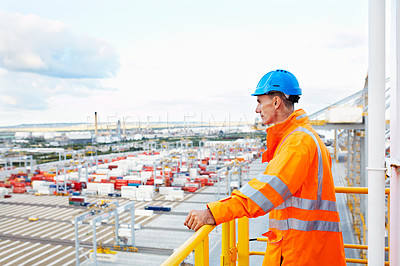 Buy stock photo Shot of a man in workwear standing on a walkway looking out over at a large commercial dock