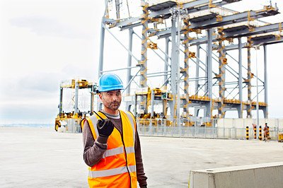 Buy stock photo Shot of a young man in workwear talking on a walkie talkie while standing on a large commercial dock