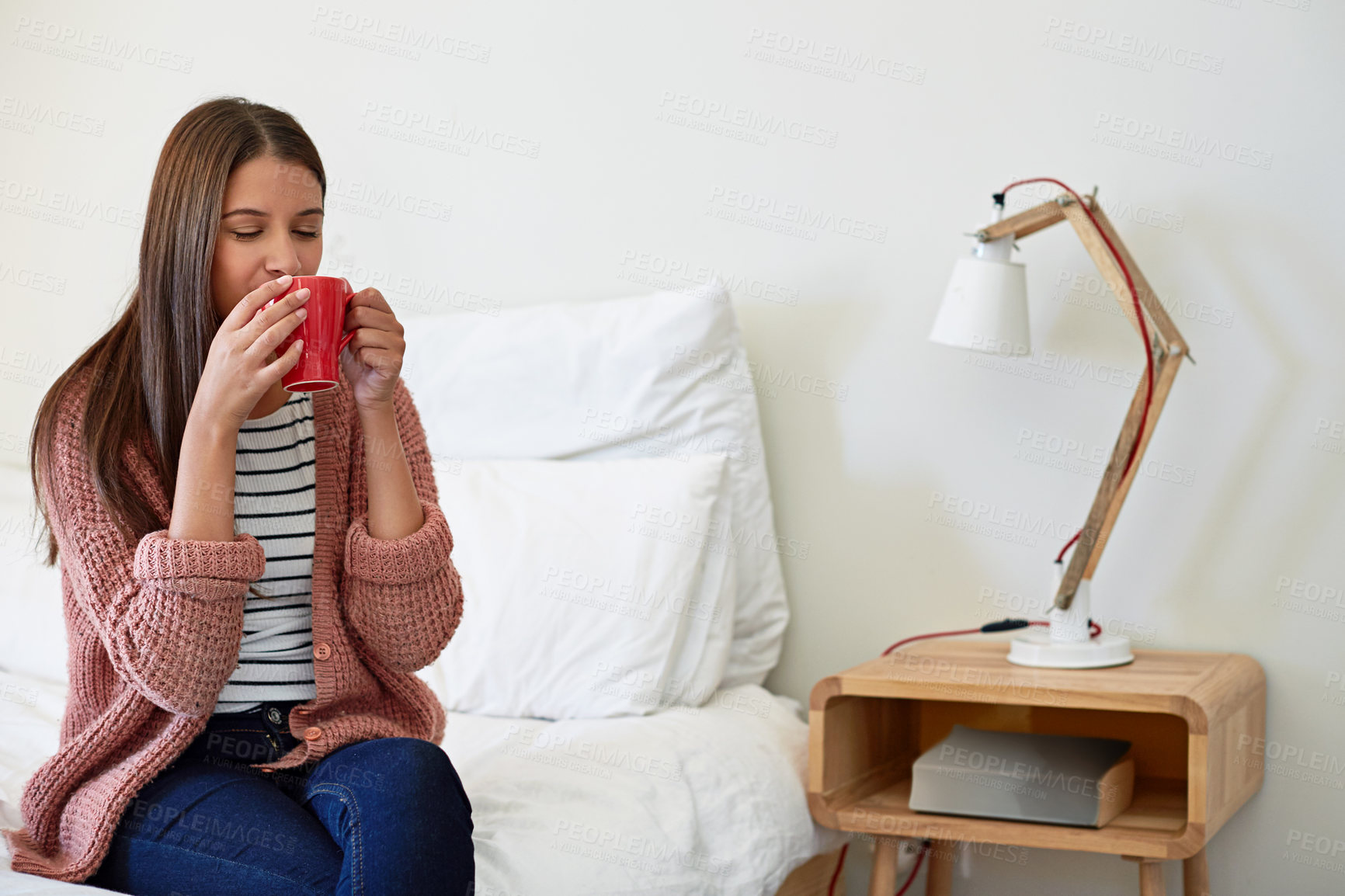 Buy stock photo Shot of a young woman sitting on her bed drinking a cup of coffee