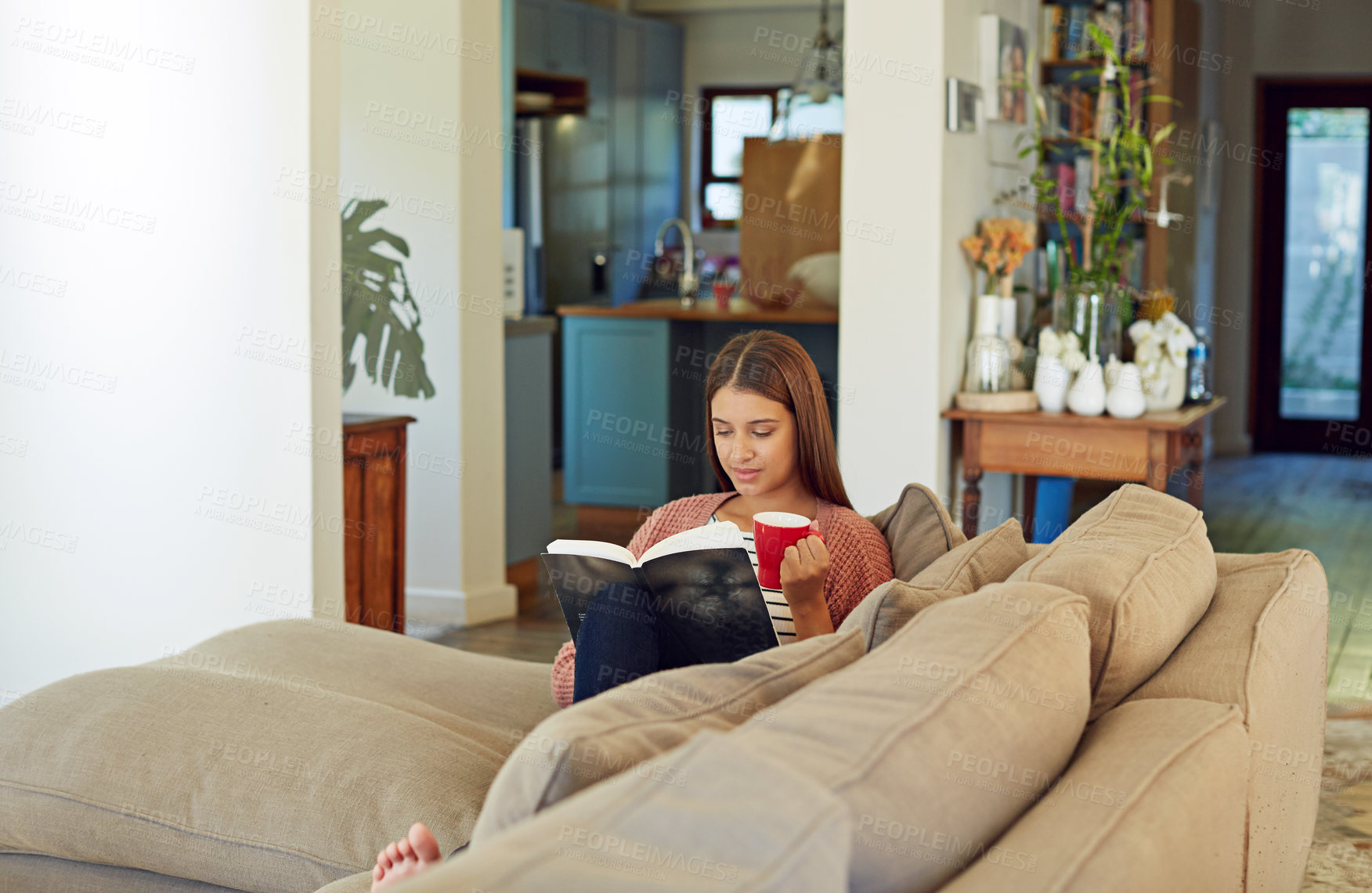 Buy stock photo Shot of a young woman sitting on her sofa reading a book and drinking a coffee