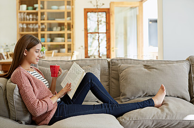 Buy stock photo Shot of a young woman sitting on her sofa reading a book and drinking a coffee