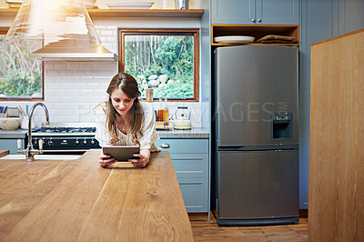 Buy stock photo Shot of a young woman leaning on her kitchen counter using a digital tablet
