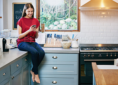 Buy stock photo Shot of a young woman sitting on her kitchen counter using a cellphone