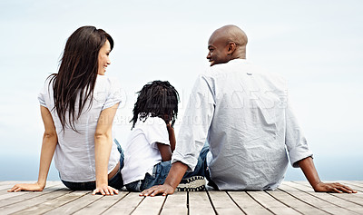 Buy stock photo Rearview shot of a young family sitting side by side on a boardwalk looking at the ocean