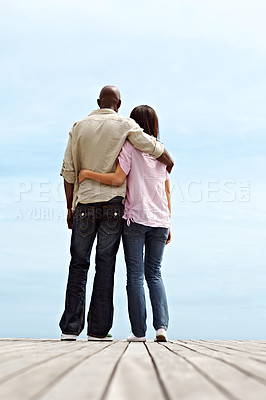 Buy stock photo Rearview shot of a young couple standing side by side on a boardwalk looking at the ocean