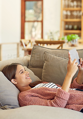 Buy stock photo Shot of a young woman lying on her sofa using a digital tablet