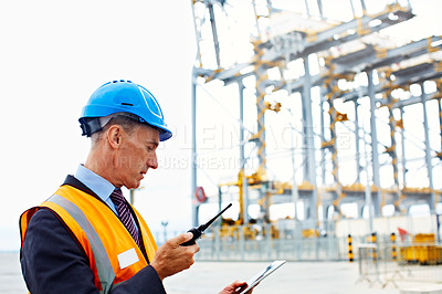 Buy stock photo Shot of a man in workwear standing outside on a large commercial dock talking on a walkie talkie