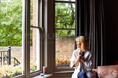 Buy stock photo Shot of a mature woman drinking tea while looking out of her living room window