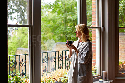 Buy stock photo Shot of a mature woman drinking tea while looking out of her living room window