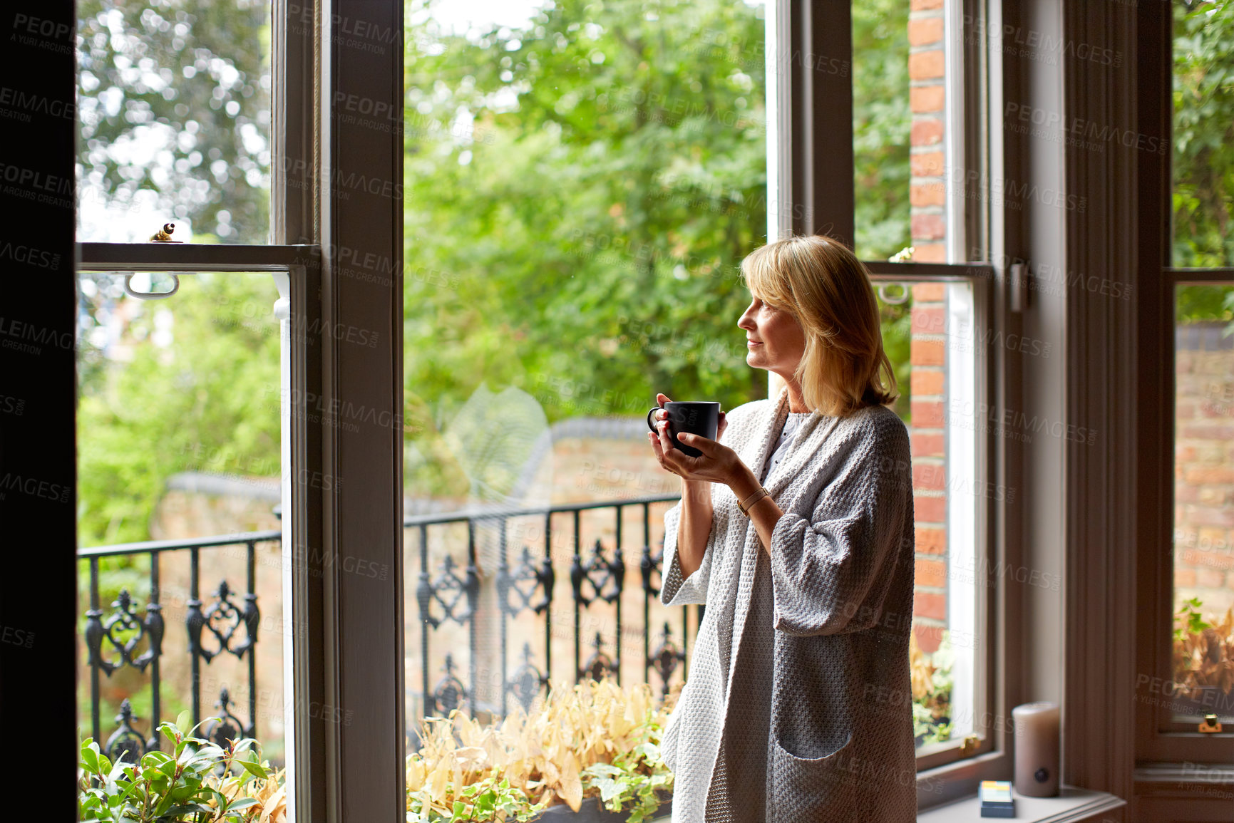 Buy stock photo Shot of a mature woman drinking tea while looking out of her living room window