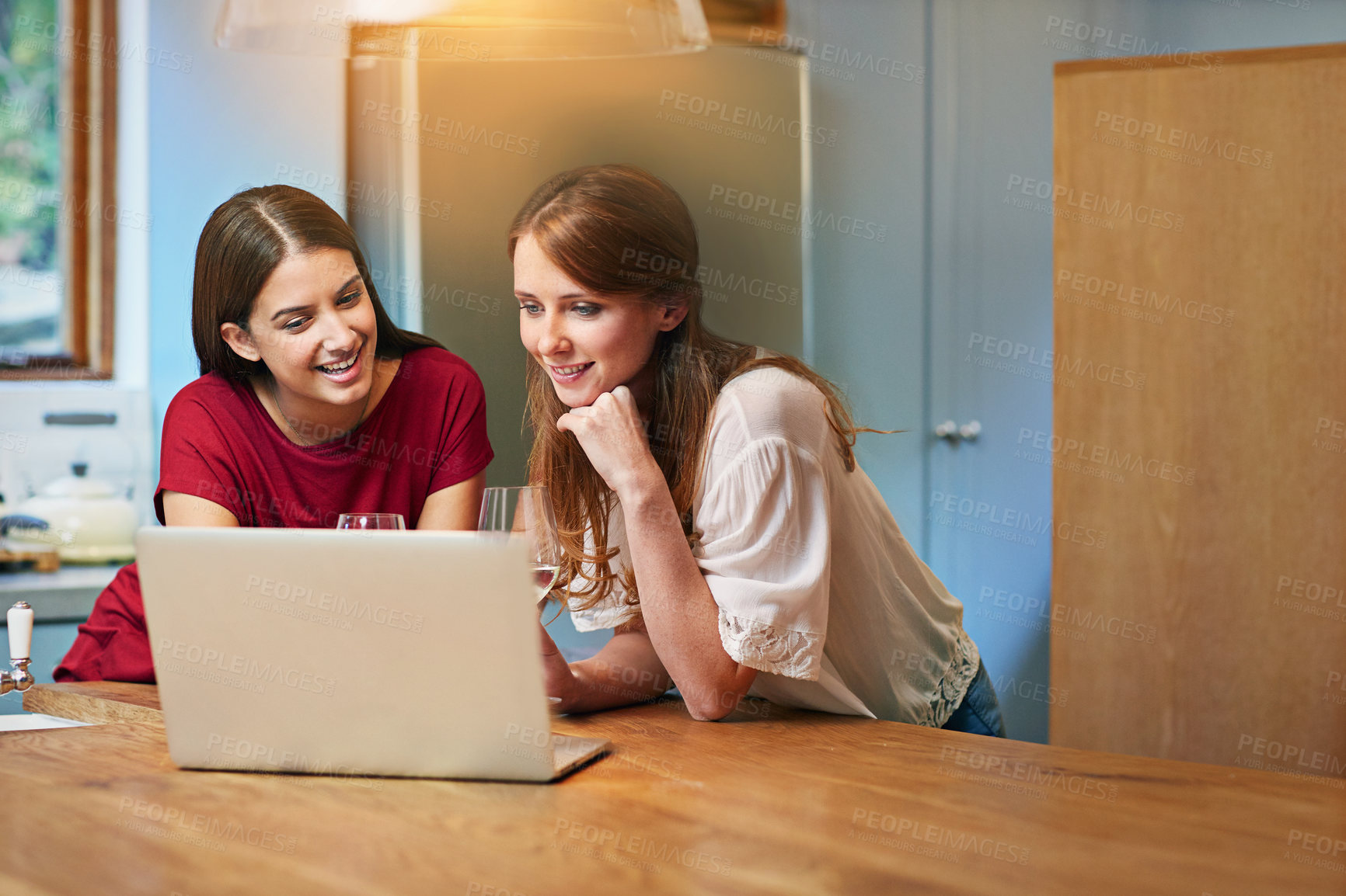 Buy stock photo Shot of two friends sitting at the kitchen table using a laptop and drinking wine