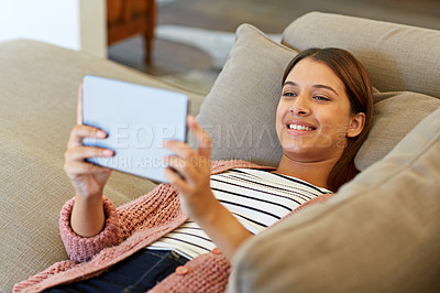 Buy stock photo Shot of a young woman lying on her sofa using a digital tablet