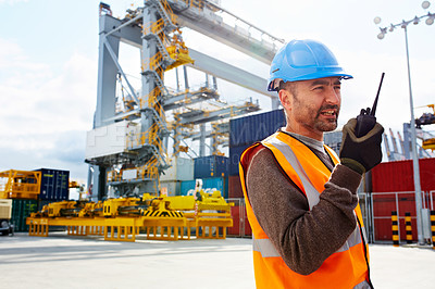 Buy stock photo Shot of a young man in workwear talking on a walkie talkie while standing on a large commercial dock